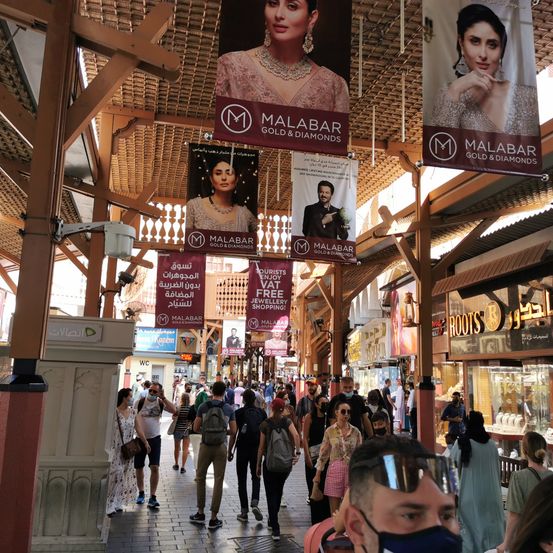 Ein geschäftiger Markt mit vielen Menschen, die herumlaufen. Werbebanner hängen von der Decke, darunter eines für Malabar Gold & Diamonds.