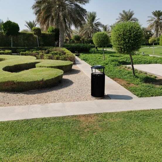 Ein gut gepflegter Gartenbereich verfügt über einen gewundenen Weg durch gepflegte Büsche, mit einem schwarzen Mülleimer an der Seite und Palmen im Hintergrund.