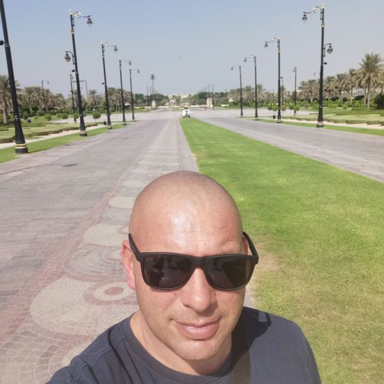 Ein kahlköpfiger Mann in Sonnenbrille steht lächelnd auf einem Bürgersteig. Dahinter befindet sich eine lange Straße mit Straßenlaternen, Gras und Bäumen.