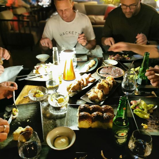 Eine Gruppe von Männern sitzt um einen Tisch mit mehreren Gerichten und Getränken, einer hält eine Gabel, der andere eine Bierflasche. Der Tisch ist mit verschiedenen Speisen bestückt, darunter Brot, Fleisch und Gemüse.