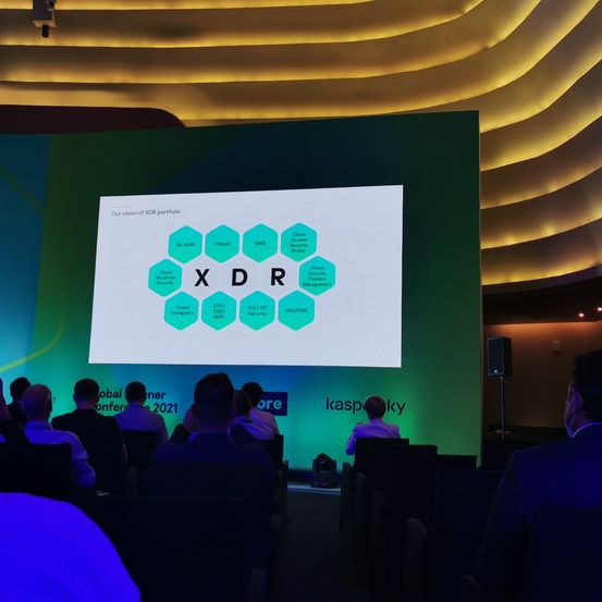 Ein Konferenzraum mit einer großen Leinwand, die eine Präsentation zeigt. Das Publikum sitzt vor der Leinwand. Die Präsentation enthält Text und Logos.