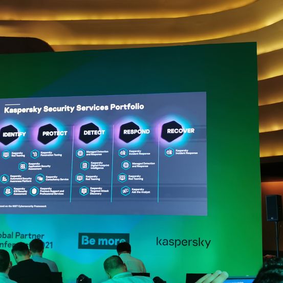 Eine Präsentationsfolie für Kaspersky Security Services Portfolio wird auf einem Bildschirm angezeigt. Es zeigt fünf Abschnitte: Identifizieren, Schützen, Erkennen, Reagieren und Wiederherstellen. Menschen sitzen davor.