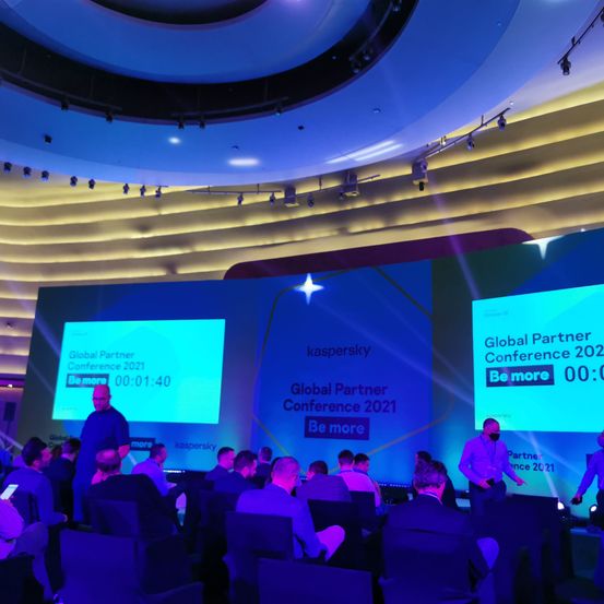 Publikum in Stühlen auf der Global Partner Conference 2021, mit einem Redner, der steht, und einer großen Leinwand, die 'Be more' und die Zeit anzeigt.