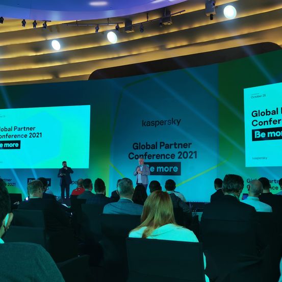 Eine Konferenz mit einer großen Leinwand, auf der 'Global Partner Conference 2021' steht. Zwei Redner stehen auf der Bühne, und viele Teilnehmer sitzen davor.