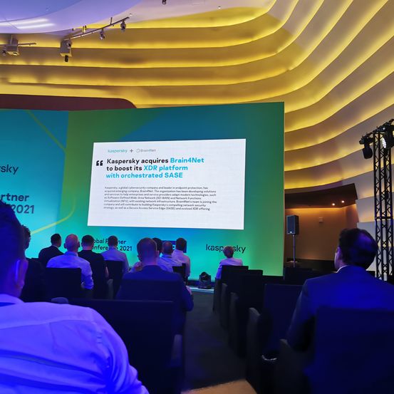 Zuhörer sitzen auf Stühlen bei einer Konferenz und hören einer Präsentation auf einer großen Leinwand zu, auf der steht 'Kaspersky erwirbt Brain4Net, um seine XDR-Plattform mit orchestriertem SASE zu stärken.'