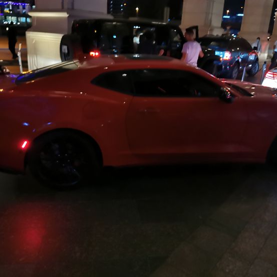 Ein roter Sportwagen steht in einem schwach beleuchteten Bereich neben anderen Fahrzeugen. Eine Person in einem weißen T-Shirt steht in der Nähe eines schwarzen SUV mit offener Tür.