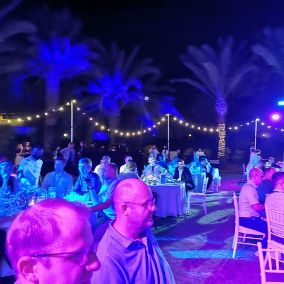 Eine nächtliche Outdoor-Veranstaltung mit Tischen und Stühlen unter blauen Lichtern. Die Leute sitzen, einige mit Brillen und Masken, mit Palmen und Lichterketten im Hintergrund.