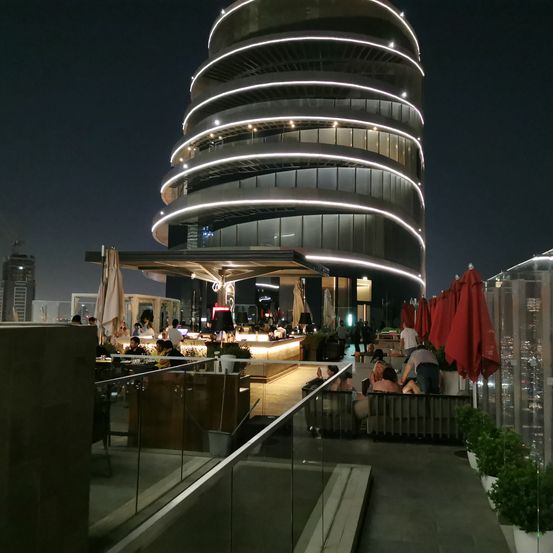 Bei Nacht ein modernes, kreisförmiges Gebäude mit vielen Glasfenstern und einer Bar. Menschen sitzen auf Sofas unter roten Sonnenschirmen und genießen Getränke.