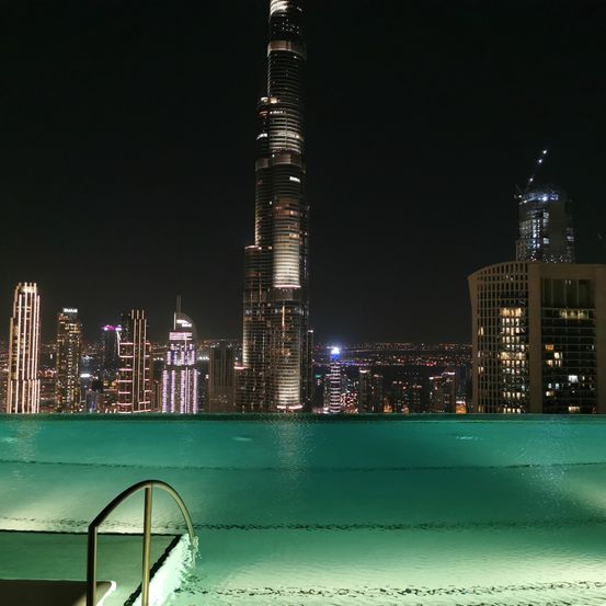 Ein Nachtblick auf die Skyline einer Stadt mit einem hohen Wolkenkratzer und anderen Gebäuden. Ein Swimmingpool ist sichtbar, mit einem Handlauf auf der linken Seite.