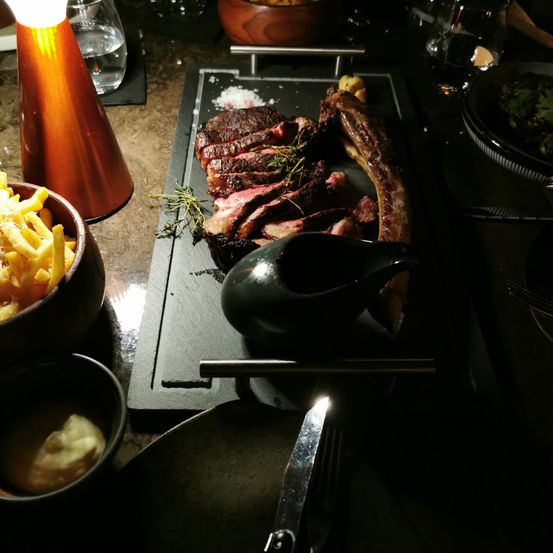Eine schwarze Schieferplatte mit Steak und einer schwarzen Gravertasse. Pommes Frites, Soße und eine Schüssel sind auf dem Tisch. Eine Lampe und ein Glas sind ebenfalls auf dem Tisch.