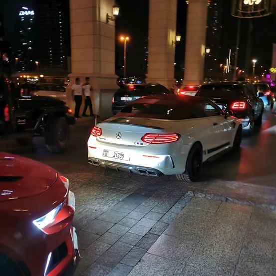 Ein weißer Mercedes Cabriolet ist nachts am Straßenrand geparkt, mit mehreren anderen Autos dahinter. Zwei Personen in weißen Hemden gehen in der Nähe vorbei.