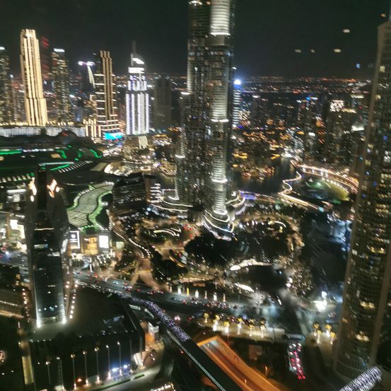 Ein nächtlicher Luftblick auf eine geschäftige Stadt mit vielen Hochhäusern, die von zahlreichen Lichtern erhellt werden, und einem Fluss, der durch die urbane Landschaft fließt.