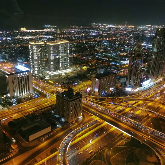 Ein nächtlicher Luftblick auf eine geschäftige Stadt mit hohen Gebäuden, einem komplexen Netzwerk von Straßen und Autobahnen und zahlreichen Lichtern, die die Landschaft erhellen. Der Himmel ist dunkel, und die Lichter der Stadt reflektieren von den Gebäuden.