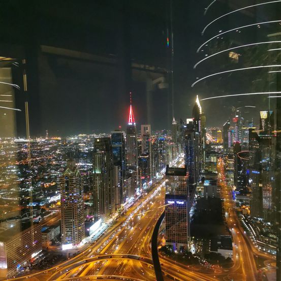 Ein Nachtblick auf eine Stadt mit vielen Hochhäusern und einer belebten Straße mit vielen Autos. Die Stadt ist von vielen Lichtern beleuchtet, darunter ein rotes Licht auf einem Turm.