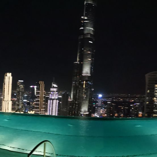 Ein Nachtblick auf eine Stadtsilhouette mit einem großen Pool im Vordergrund, beleuchteten Gebäuden im Hintergrund und einem hohen Wolkenkratzer in der Mitte.