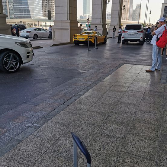 Ein gelbes Sportauto ist auf einem Parkplatz in der Stadt geparkt. Mehrere Leute stehen herum und bewundern das Auto. Eine Person mit einer roten Tasche macht ein Foto.