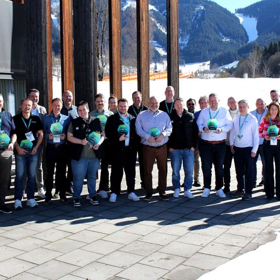 Eine Gruppe von Menschen posiert für ein Foto mit Auszeichnungen in einem verschneiten Berggebiet. Jeder hält eine grüne Auszeichnung und steht vor einer Holzstruktur.