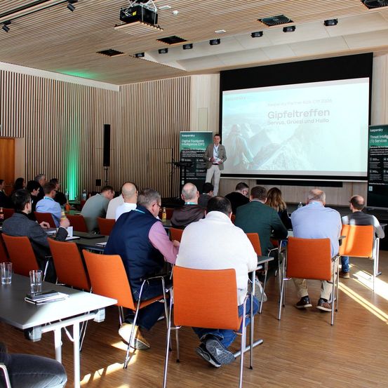 Eine Gruppe von Menschen nimmt an einer Präsentation in einem Konferenzsaal teil. Ein Mann spricht vorne, steht vor einer Leinwand mit 'Gipfeltreffen'.