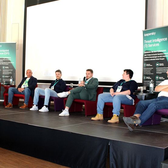 Fünf Männer sitzen auf einer Bühne, wahrscheinlich während einer Konferenz. Ein Mann hält ein Mikrofon. Ein Banner mit dem Text 'kaspersky Threat Intelligence (TI) Services' ist hinter ihnen zu sehen.