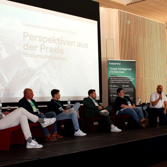 Fünf Männer sitzen auf einer Bühne, wahrscheinlich auf einer Konferenz, und tragen Turnschuhe. Einer hält ein Mikrofon. Eine Projektionsleinwand zeigt eine Präsentation mit dem Titel 'Kaspersky Partner Kick-Off 2026 Perspektiven aus der Praxis Podiumsdiskussion.'