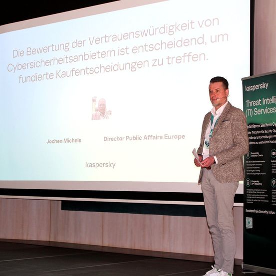 Ein Mann steht auf der Bühne und präsentiert vor einer Projektionsleinwand, auf der steht: 'Die Bewertung der Vertrauenswürdigkeit von Cybersicherheitsanbietern ist entscheidend, um fundierte Kaufentscheidungen zu treffen.'