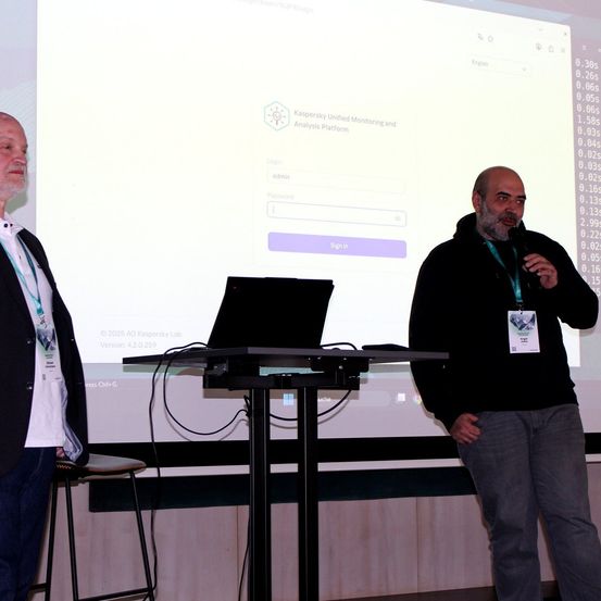 Zwei Männer stehen vor einer Projektionsleinwand, die die Kaspersky Unified Monitoring and Analysis Platform zeigt. Ein Mann spricht in ein Mikrofon, während der andere auf den Bildschirm schaut. Ein Laptop steht auf dem Podium.