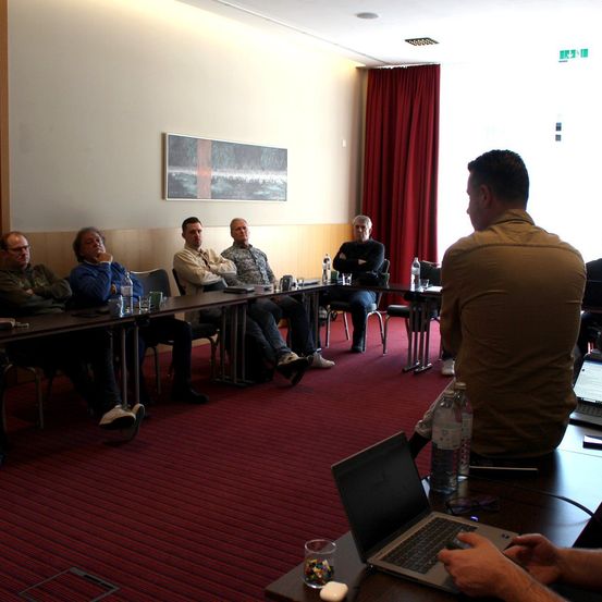 Eine Gruppe von Männern sitzt um einen Tisch in einem Konferenzraum. Flaschen, Tassen und Laptops stehen auf dem Tisch. Ein Mann steht vorne und führt wahrscheinlich eine Besprechung. Ein Gemälde hängt an der Wand.
