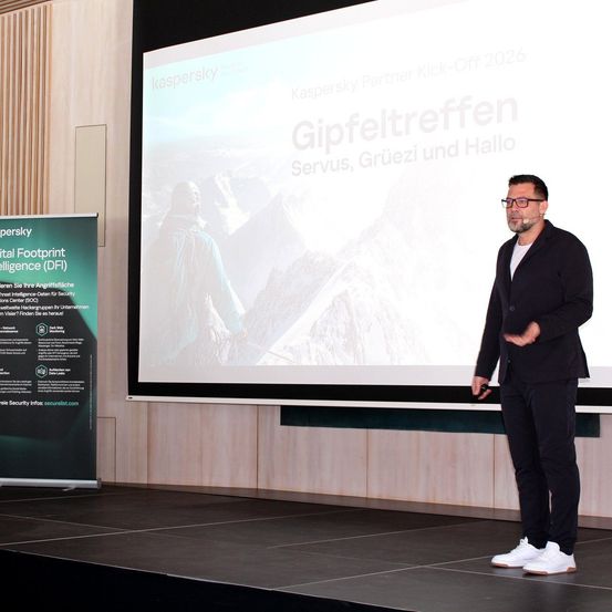 Ein Mann auf der Bühne, der bei einer Veranstaltung präsentiert, trägt eine Brille und Turnschuhe. Hinter ihm zeigt eine große Leinwand ein Bild eines Mannes und den Text 'Gipfeltreffen'. Ein Banner befindet sich auf der linken Seite der Bühne.
