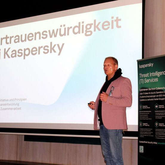 Ein Mann steht vor einer Leinwand, auf der 'rtrauenswürdigkeit i Kaspersky' steht. Er hält ein Handy in der Hand und scheint eine Präsentation zu halten.