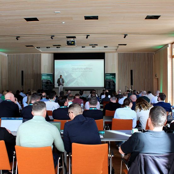 Ein Konferenzsaal mit einer Projektionsleinwand, auf der 'Gipfeltreffen' steht. Ein Mann spricht auf der Bühne, während zuhörende Teilnehmer in formeller Kleidung zuhören. Der Raum ist mit orangefarbenen Stühlen und Tischen gefüllt.