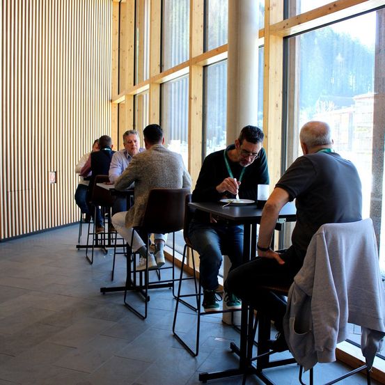 Einige Männer sitzen an Tischen in einem Café. Das Interieur besteht aus Holzwänden und großen Fenstern. Ein Mann schreibt.