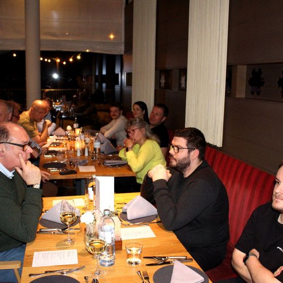 Eine Gruppe von Menschen sitzt in einem schwach beleuchteten Restaurant an Tischen, mit Weingläsern, Tellern und Besteck auf dem Tisch. Der Mann in Schwarz schaut nach unten, während der Mann in Grün sein Kinn berührt.