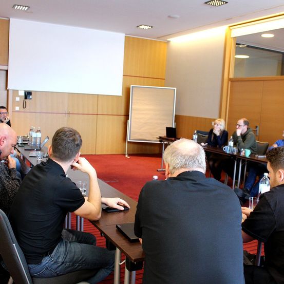 Ein Besprechungsraum mit rotem Teppich zeigt Menschen in schwarzen Shirts, die um Tische herumsitzen. Ein Whiteboard und eine Leinwand sind im Hintergrund zu sehen.