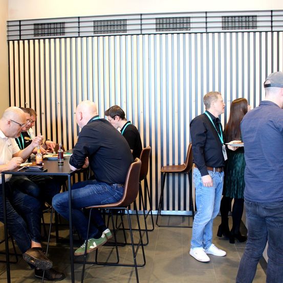 Eine Gruppe von Menschen in einem Besprechungsraum isst. Ein Mann steht und redet. Andere sitzen und essen.