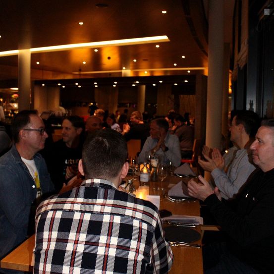 Eine Gruppe von Menschen sitzt in einem Restaurant an einem Tisch, einige klatschen, während andere Getränke und Essen auf ihren Tischen haben.