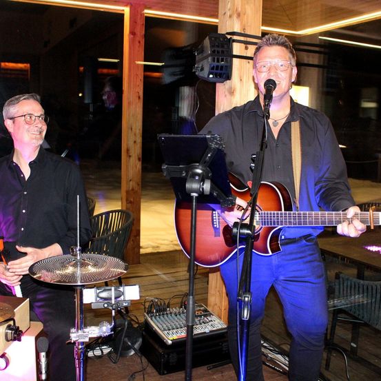 Zwei Männer spielen auf einer Bühne; einer spielt Gitarre und singt in ein Mikrofon, während der andere Schlagzeug spielt. Der Raum ist mit Holzböden und Glaswänden ausgestattet.