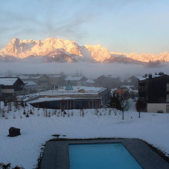 Eine verschneite Landschaft mit einem Pool, umgeben von Gebäuden und Bäumen. Im Hintergrund sind verschneite Berge unter einem blauen Himmel sichtbar.