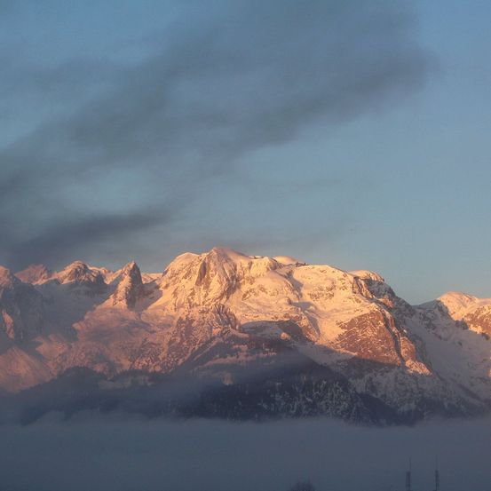 Ein Gebirgszug ist unter einem bewölkten Himmel von Schnee bedeckt. Die Sonne geht unter und taucht die Landschaft in ein warmes Licht. Nebel umgibt den unteren Teil der Berge.