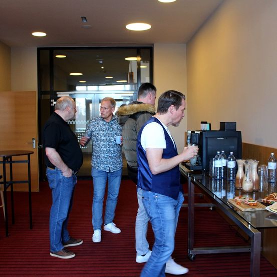 Vier Männer stehen in einem Raum in der Nähe eines Tisches mit Erfrischungen. Ein Mann hält einen Becher. Eine Tür ist hinter ihnen.