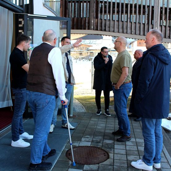 Eine Gruppe von Männern steht im Winter vor einem Gebäude. Ein Mann zeigt, ein anderer hält einen Stock, und die anderen schauen zu. Schnee bedeckt den Boden.