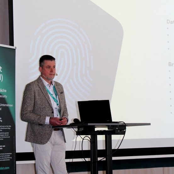 Ein Mann steht vor einer Projektionswand mit Laptop und hält eine Präsentation. Er trägt ein Mikrofon und ein Geschäftsoutfit.