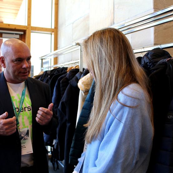 Ein Mann mit einem grünen Ausweisband spricht mit einer Frau in einem hellblauen Pullover, beide stehen vor einem Ständer mit schwarzen Mänteln.