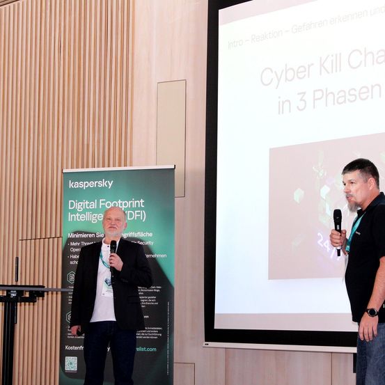 Zwei Männer sprechen in Mikrofone vor einer Leinwand mit einer Präsentation mit dem Titel 'Cyber Kill Chain in 3 Phasen.' Ein Banner mit 'Digital Footprint Intelligence (DFI)' steht dahinter.