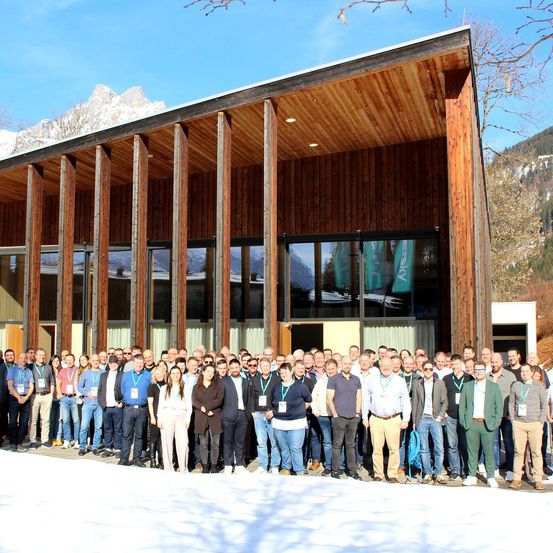 Eine Gruppe von Menschen in formeller Kleidung posiert vor einem modernen Gebäude mit Glaswänden und Holzakzenten. Im Hintergrund sind schneebedeckte Berge zu sehen.