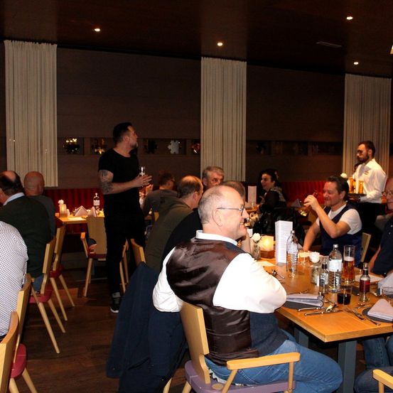 Eine Gruppe von Menschen versammelt sich in einem Restaurant und trinkt Bier. Ein Mann steht und spricht, während andere zuhören. Der Raum hat Holzböden, rote Wände und weiße Vorhänge.