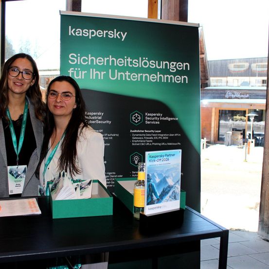 Zwei Frauen stehen lächelnd hinter einem Tisch, neben einem grünen Banner mit Kaspersky-Marke. Das Banner trägt die Aufschrift 'Sicherheitslösungen für Ihr Unternehmen' und zeigt verschiedene Sicherheitsdienste.