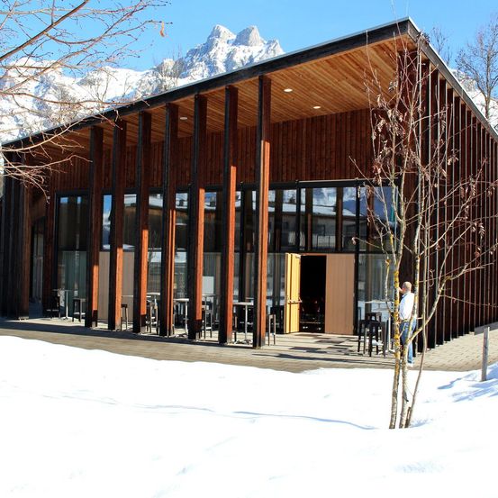 Ein modernes Holzgebäude mit Glaswänden steht vor einem verschneiten Berg. Ein Mann steht draußen und schaut sich etwas an.