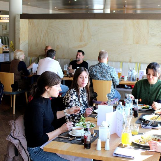 Ein Restaurant mit Gästen, die an Tischen essen, einige im Gespräch, andere konzentrieren sich auf ihr Essen. Verschiedene Teller, Gläser und Beilagen sind auf dem Tisch.