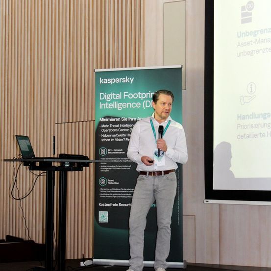 Ein Mann hält eine Präsentation vor einer Projektionsleinwand mit einem Banner über digitale Fußabdruck-Intelligenz. Er hält ein Mikrofon in der Hand und trägt einen Gürtel.