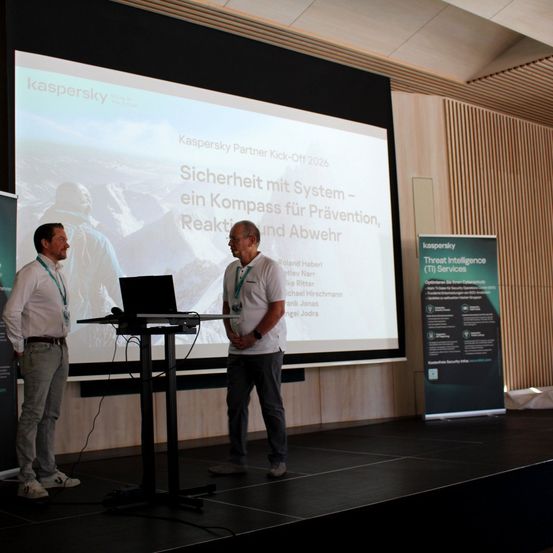 Zwei Männer stehen auf der Bühne vor einer großen Leinwand, auf der eine Kaspersky-Präsentation zu sehen ist. Der Titel der Präsentation lautet 'Sicherheit mit System - ein Kompass für Prävention, Reaktion und Abwehr.'